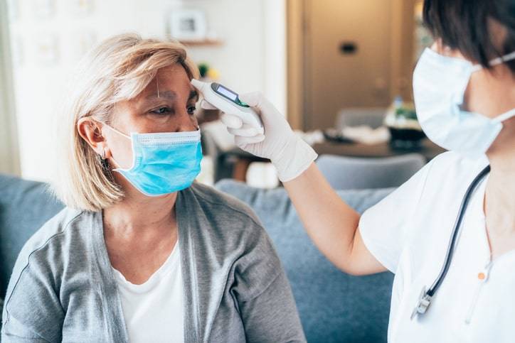A healthcare worker checks the temperature of a seated woman using a forehead thermometer in a home setting. Both are wearing masks, creating a reassuring and professional atmosphere reminiscent of a walk-in clinic.