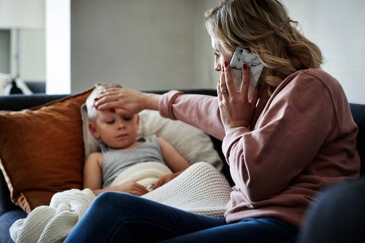 A woman is sitting on a couch, holding a phone to her ear with one hand while placing her other hand on the forehead of a sick child lying beside her, likely calling the medical center for assistance.