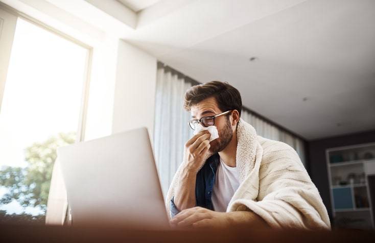 A man sitting at a table with a laptop, covering his nose with a tissue while wrapped in a blanket, looks like he might need urgent care.