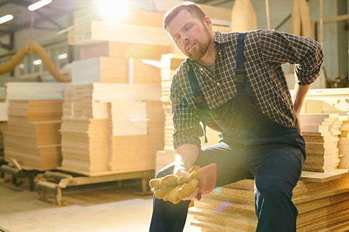 Man in work overalls and gloves sits on a stack of wood panels in a workshop, holding his lower back with a pained expression, considering whether he should visit an urgent care center.