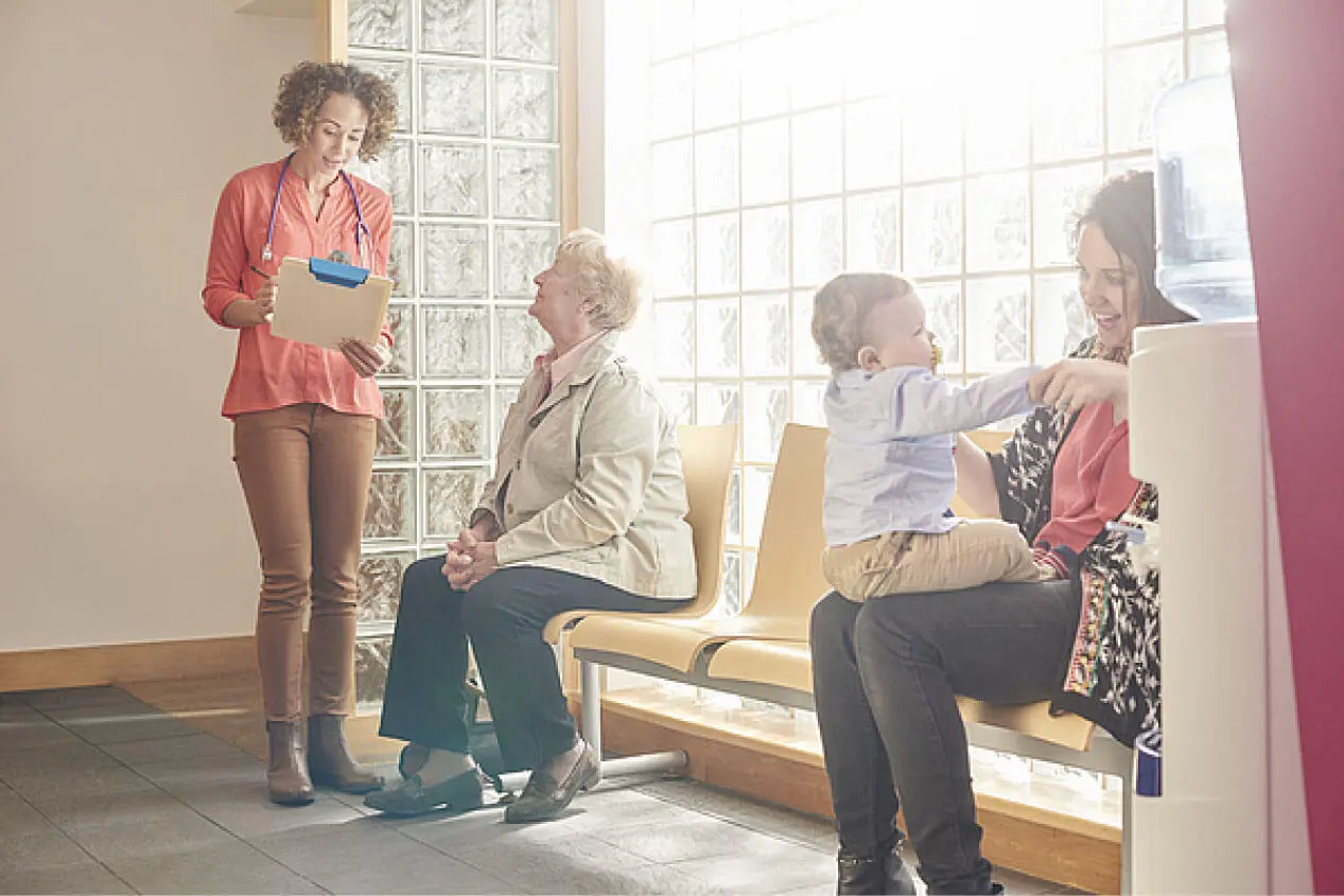 Waiting room with four patients - a mother playing with her child sitting down, and a young lady medical professional talking with a seated patient waiting in the room