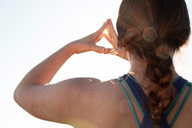 Person with a braid, seen from behind, holding hands in a triangle shape against the bright sunlight, wearing a sleeveless top near a walk-in clinic.