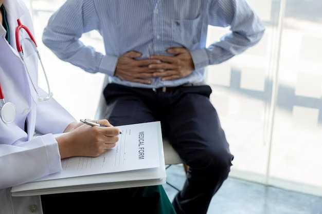 A doctor takes notes at the medical center while a patient sits nearby holding his stomach in discomfort.