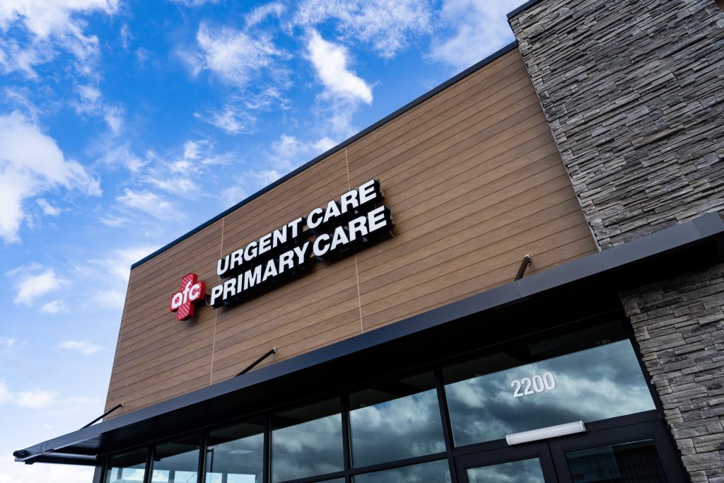 Exterior of a building with a sign reading "AFC Urgent Care Primary Care" and the number "2200" near the entrance. The modern design features wood and stone facade elements, highlighting its role as a walk-in clinic under American Family Care.