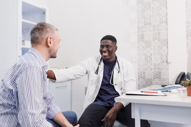 A doctor in a white coat and stethoscope is smiling and talking with a seated patient in a walk-in clinic room.