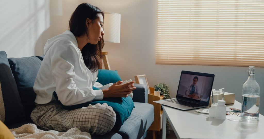 Woman sitting on a sofa, participating in a video call with an urgent care on a laptop. A water bottle, medication, and tissues are on the table beside her. Sunlight filters through blinds.