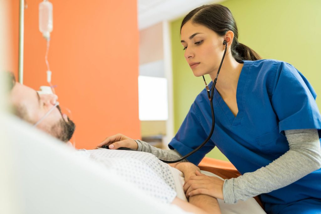 A healthcare worker in blue scrubs uses a stethoscope to check the vital signs of a patient lying in a hospital bed at the medical center.