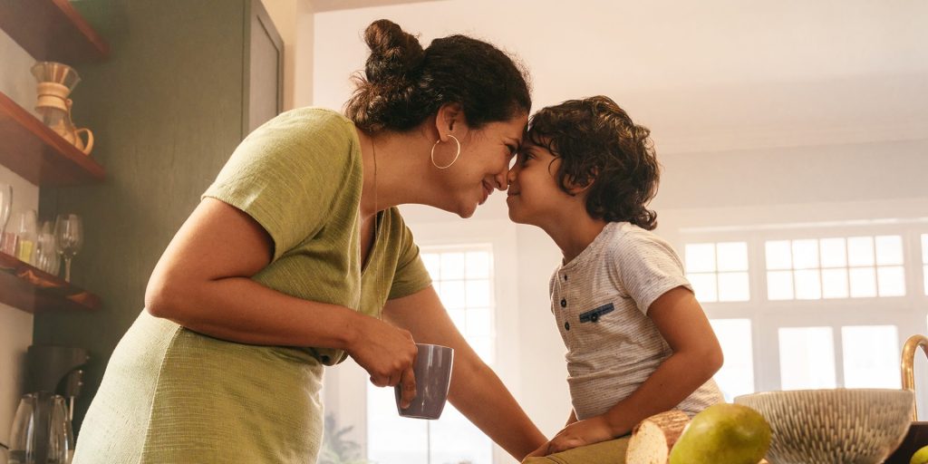 A woman and a young child touch foreheads affectionately in a kitchen. The woman holds a mug, with various kitchen items visible in the background, making it a cozy moment before heading out for their annual physicals.