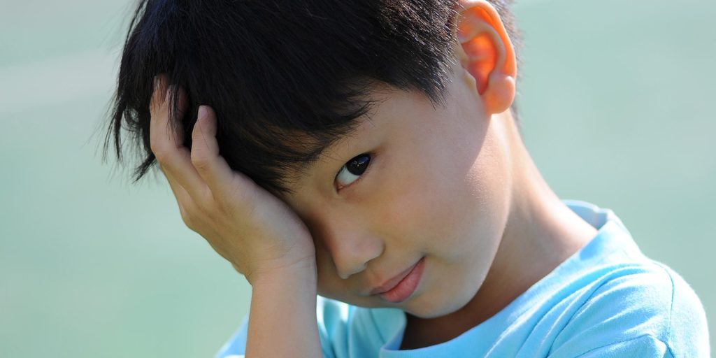A child with short dark hair wearing a light blue shirt holds their hand to their forehead, gazing directly at the camera with a slight smile, perhaps waiting at the medical center for a routine physical.