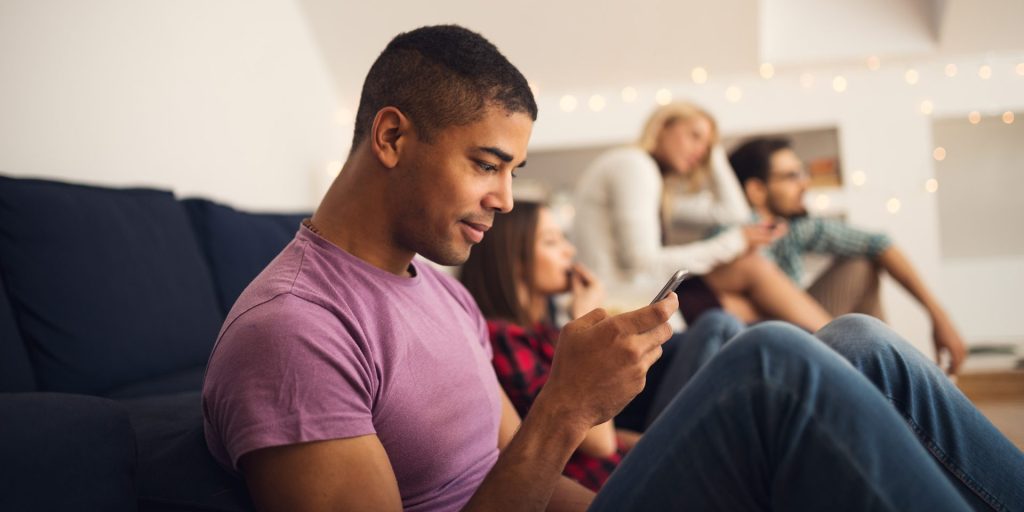 A man sits on a couch using a smartphone, possibly scheduling his next physical, while others sit in the background with one person also looking at a device.