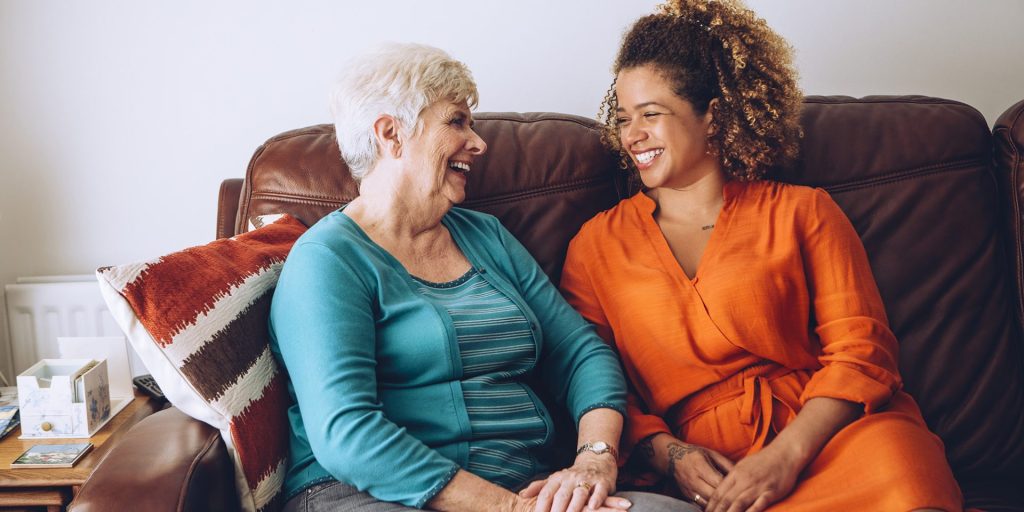 An older woman and a younger woman sit on a couch at the walk-in clinic, smiling and looking at each other.