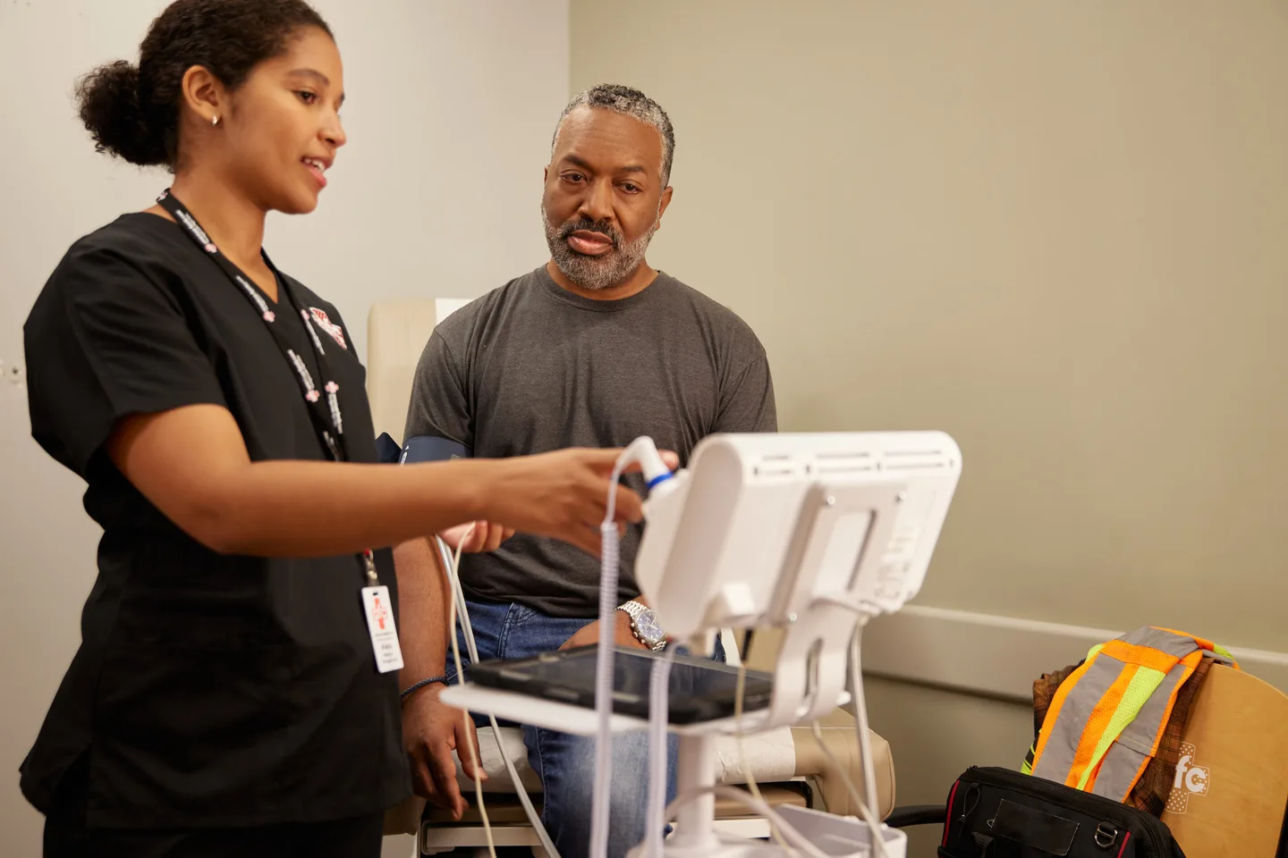 A nurse in a black uniform uses medical equipment while performing lab testing as a patient in a gray shirt, seated, observes.