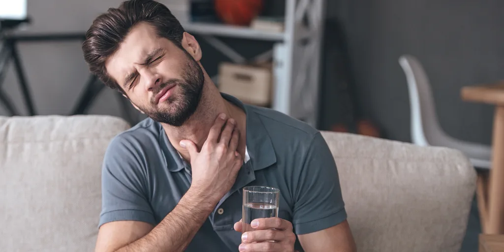 A man in a polo shirt sits on a couch, holding a glass of water in one hand and touching his throat with a pained expression, contemplating whether he should visit an urgent care clinic for relief.