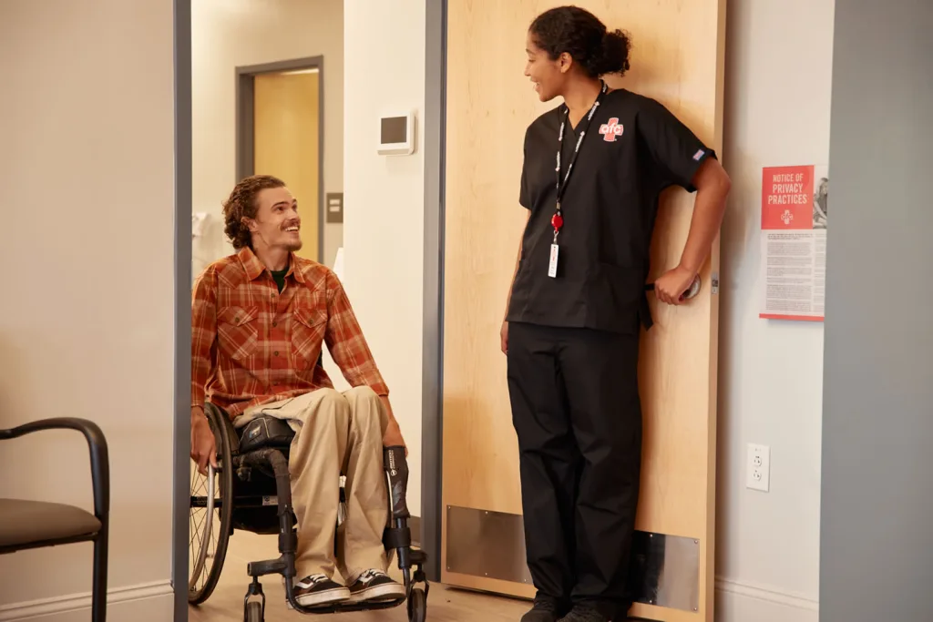 A person in a wheelchair smiles while a healthcare worker holds a door open for them. Both are engaged in conversation in a medical center setting.