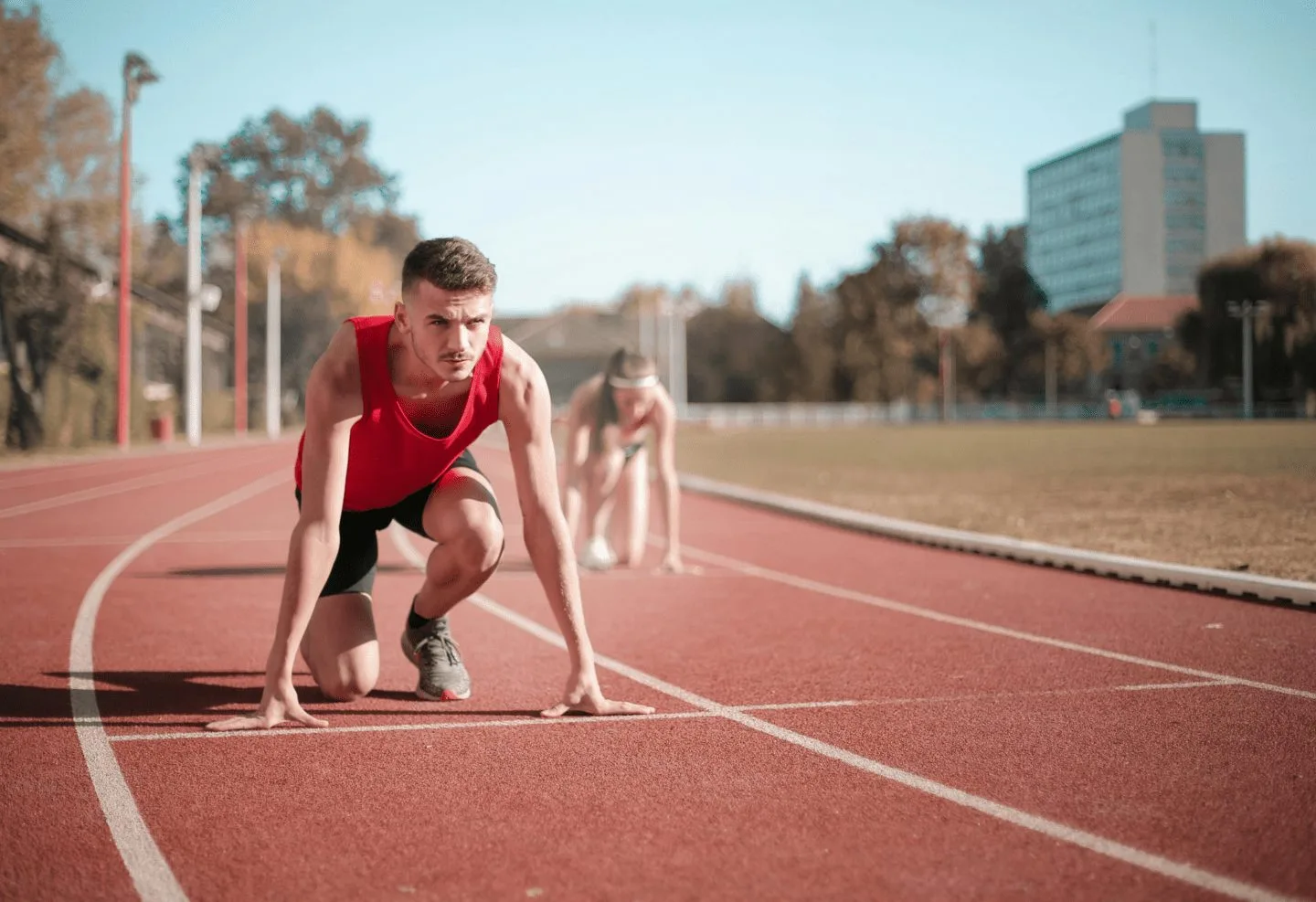 A male runner in a red tank top is crouched at the starting line on a track near the medical center, ready to race. Another runner is visible in the background, also preparing to start.