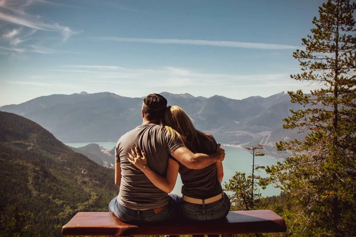 Two people sit on a bench overlooking a scenic mountainous landscape with a body of water, surrounded by trees, enjoying a peaceful moment after their visit to the nearby medical center for their annual physicals.