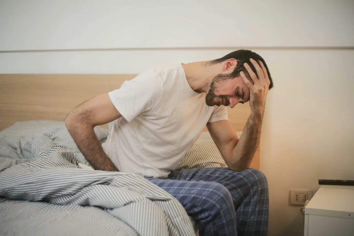 A man in a white t-shirt and plaid pajama pants sits on a bed, holding his head with his right hand and seemingly distressed. He appears to be considering whether he needs urgent care for whatever is troubling him.