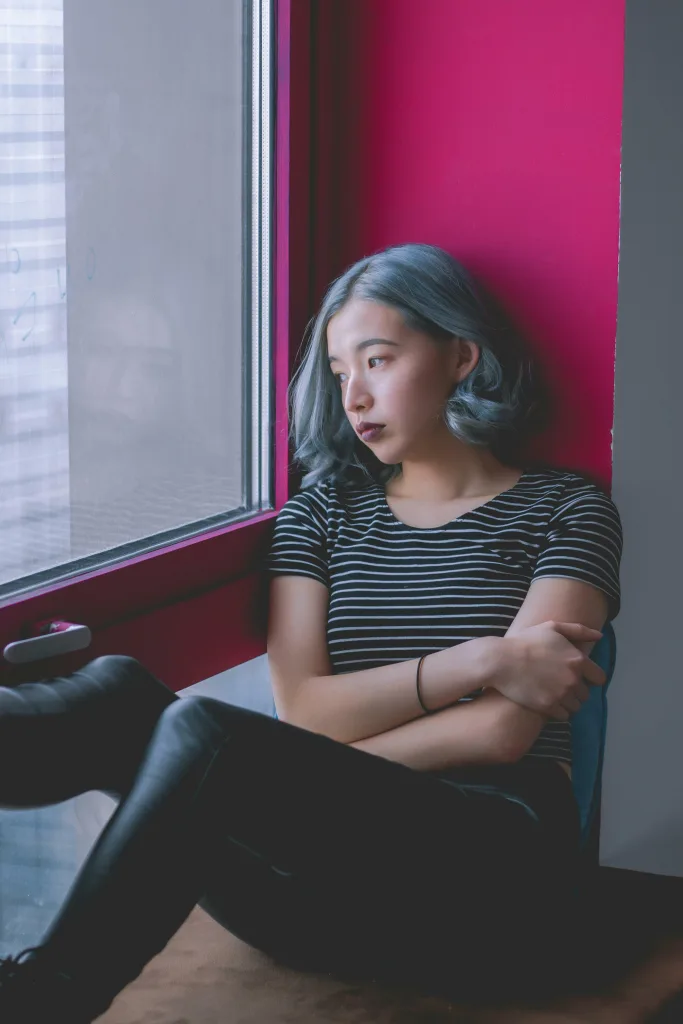 A person with blue-gray hair sits by a window, arms crossed, looking outside thoughtfully. They are wearing a black and white striped shirt and black pants, evoking the calm atmosphere of a medical center waiting room where immediate care is on hand.