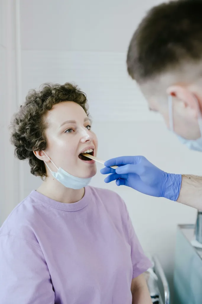 A healthcare professional in blue gloves performs a throat swab on a person wearing a light purple shirt and a partially removed face mask at an urgent care clinic.