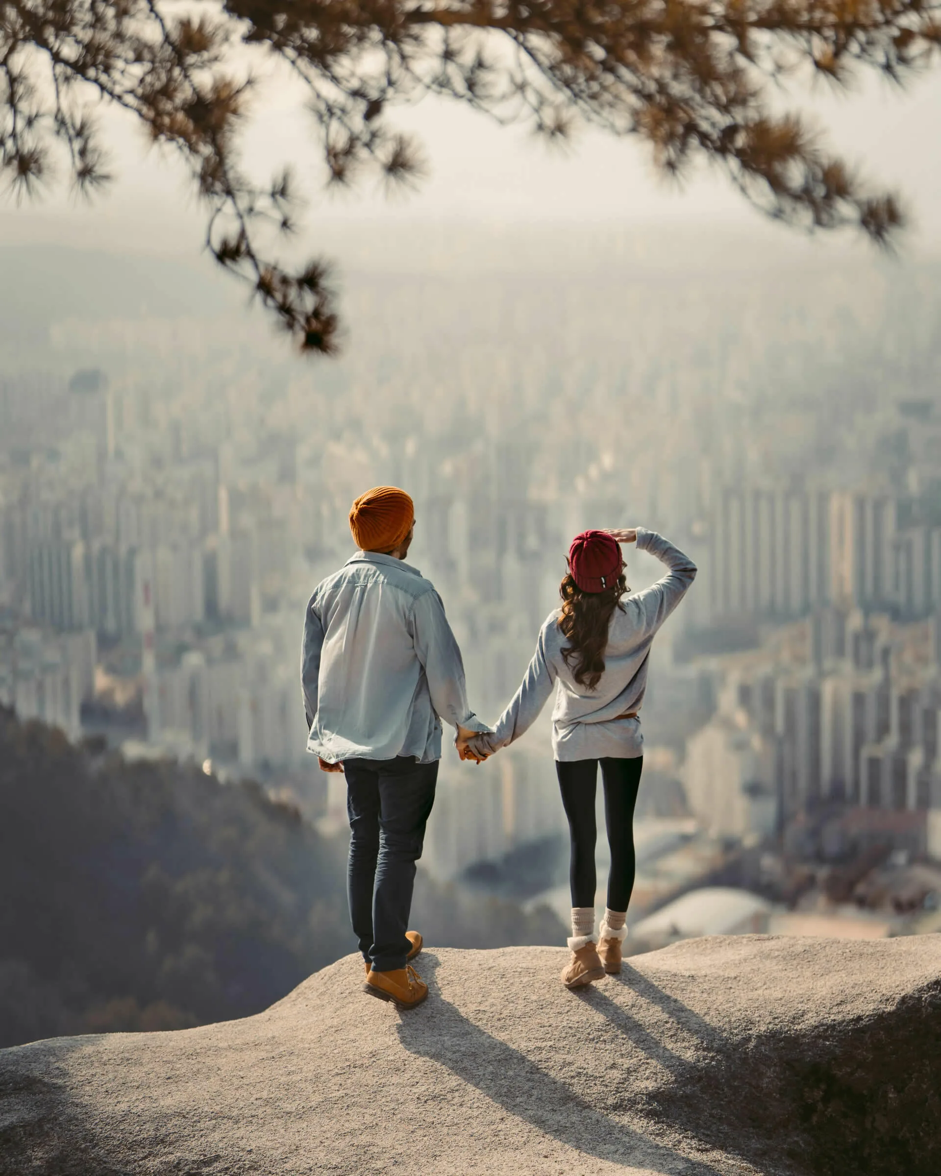 A couple holds hands while standing on a rocky cliff, overlooking a city landscape with high-rise buildings including a nearby medical center, under a tree branch.