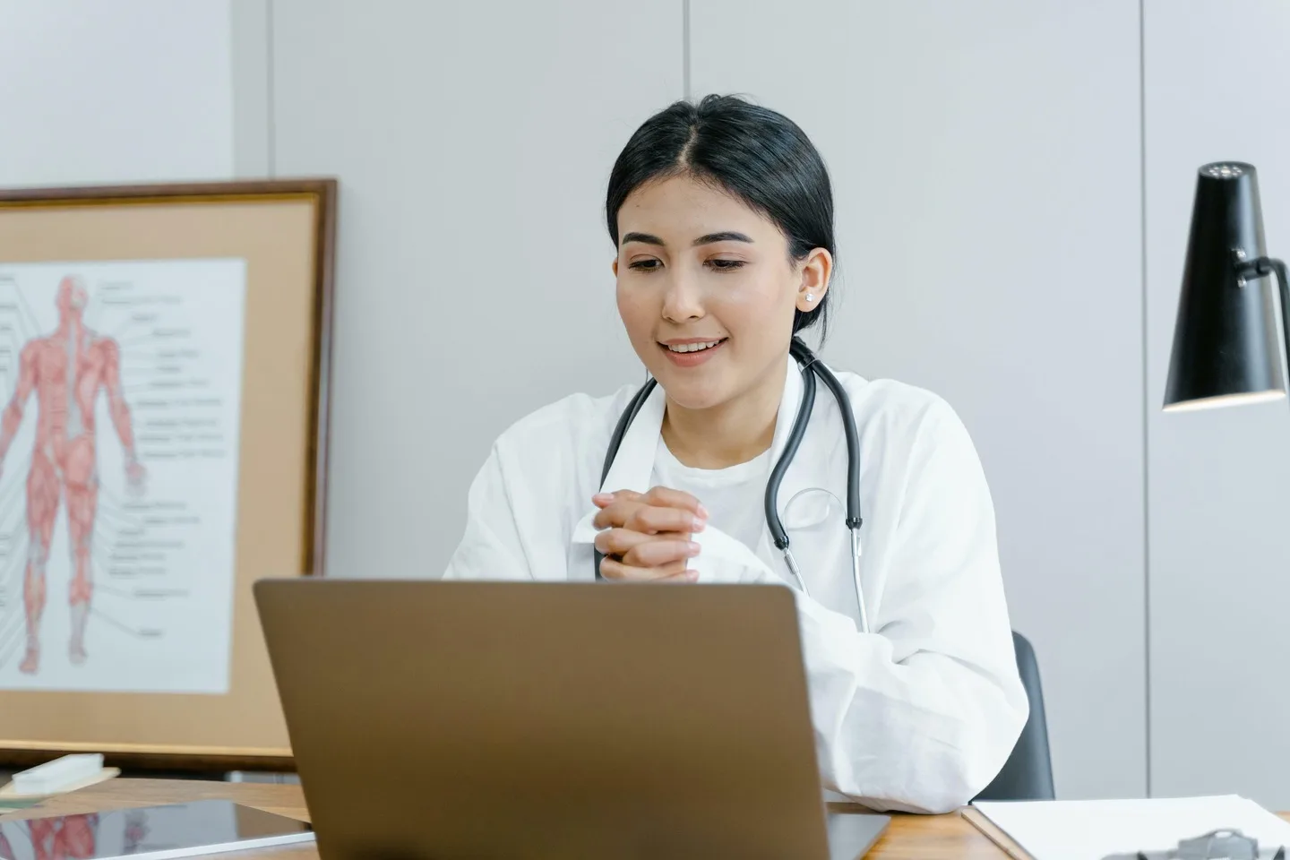 A doctor wearing a white coat and stethoscope sits at a desk with folded hands, looking at a laptop. A medical chart is visible in the background, suggesting the efficient atmosphere of an immediate care walk-in clinic.