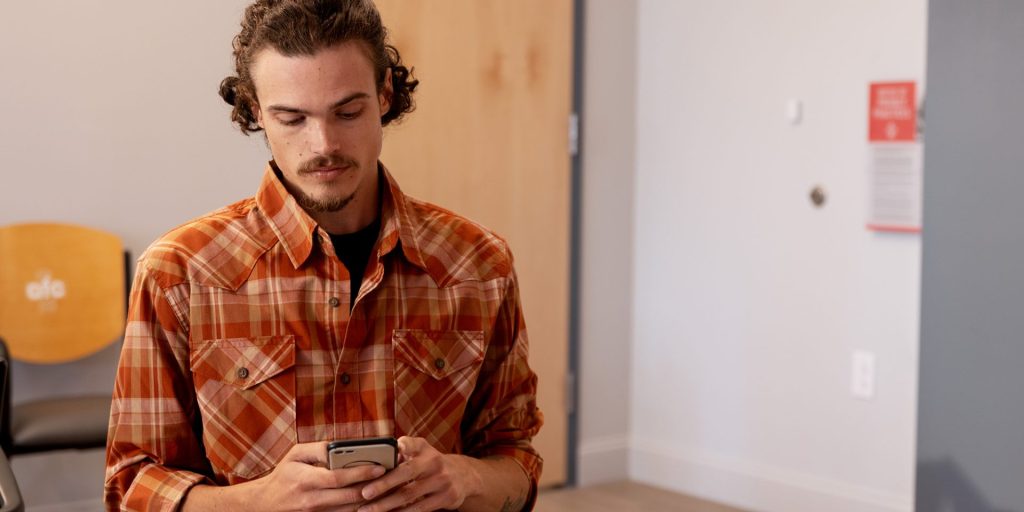 A person with curly hair and a beard, wearing an orange plaid shirt, is looking down at a smartphone in their hands. They are indoors with beige walls and a wooden door in the background, possibly waiting for lab testing results at the medical center.