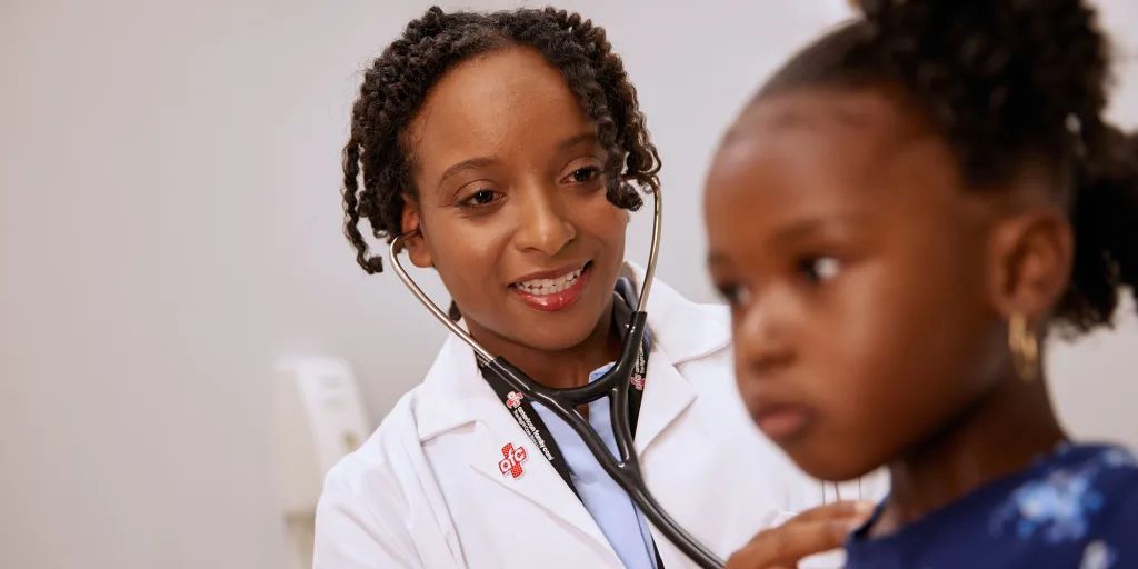 A doctor wearing a white coat and using a stethoscope examines a young girl in an immediate care setting.