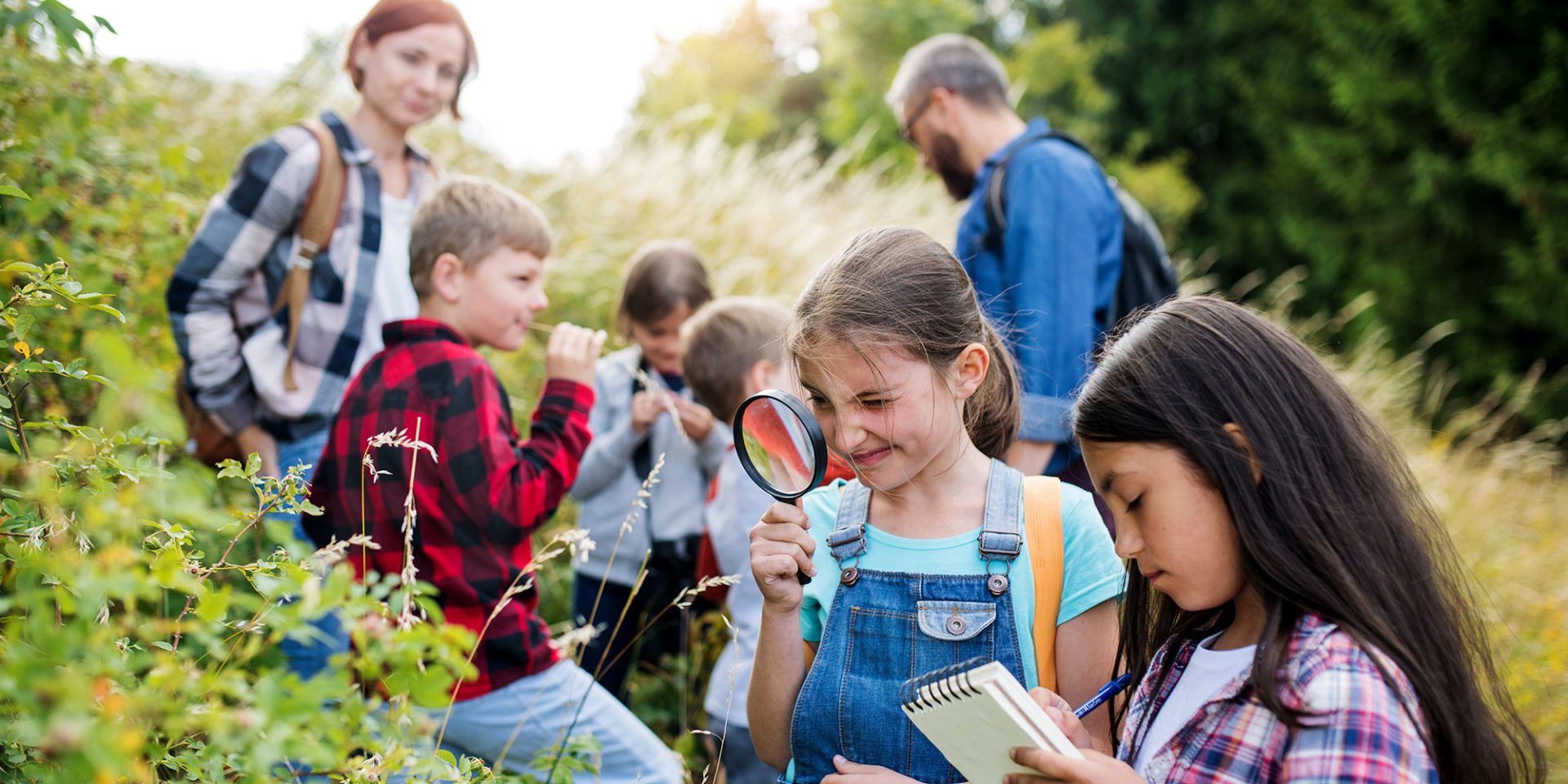 Children exploring nature with notebooks and magnifying glasses, accompanied by two adults in an outdoor setting with greenery, near a walk-in clinic.