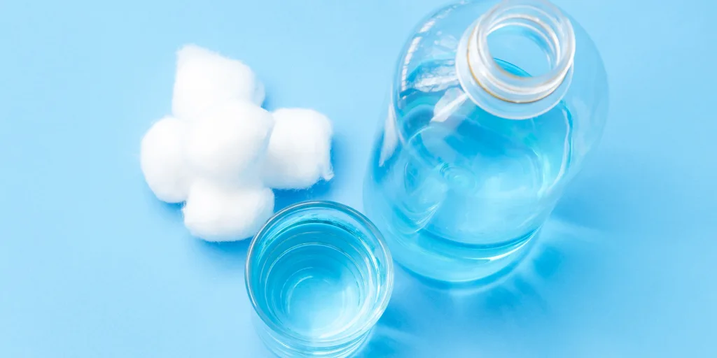 A bottle of clear liquid with an open cap, a filled glass, and four cotton balls on a blue background, reminiscent of essentials found at an urgent care clinic.