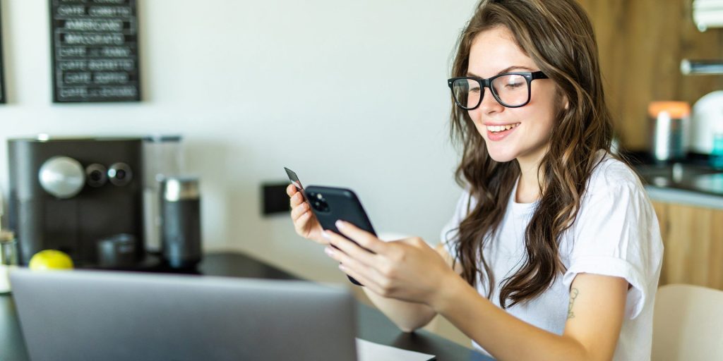 A person with glasses smiles at a smartphone while holding a credit card, seated at a table with a laptop in a kitchen, possibly scheduling an appointment at the nearby walk-in clinic.