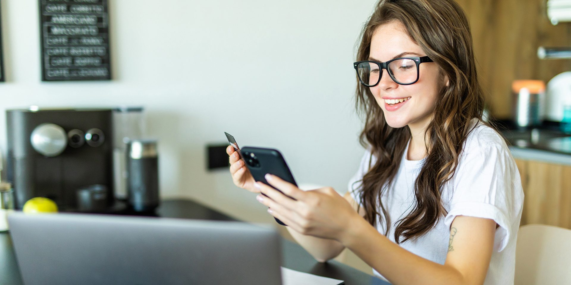A person with glasses smiles at a smartphone while holding a credit card, seated at a table with a laptop in a kitchen, possibly scheduling an appointment at the nearby walk-in clinic.