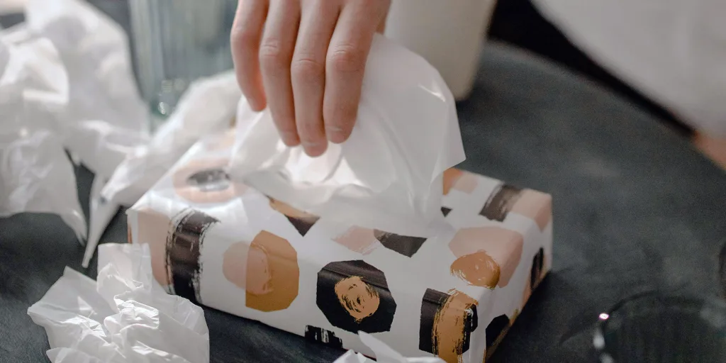 A hand reaches for a tissue from a patterned tissue box, likely found in an urgent care facility. Crumpled tissues are scattered on a dark surface around the box, hinting at a busy day addressing patient needs.