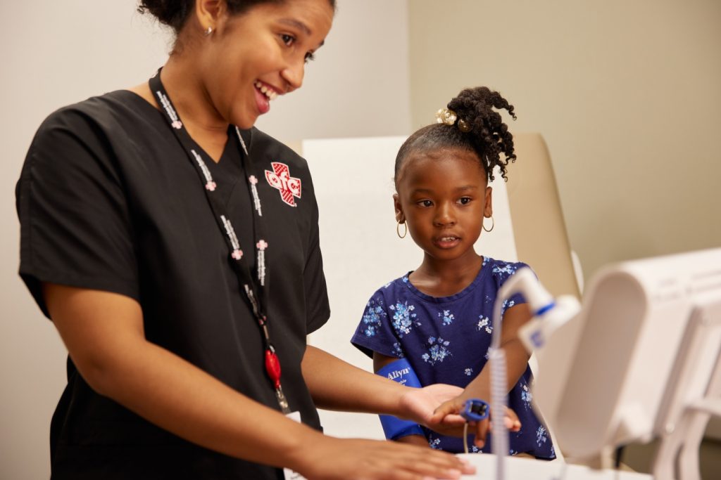 In a clinical setting at the medical center, a healthcare worker in a black uniform assists a young girl with a blood pressure monitor.