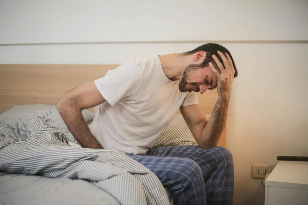 A man sits on a bed in the medical center, wearing a white shirt and checkered pajama pants, holding his head with a pained expression.