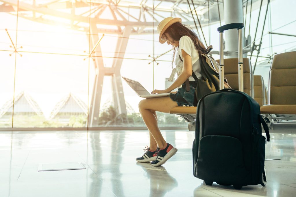 A person seated in an airport terminal, working on a laptop with a backpack beside them and a bright, sunlit glass structure in the background.