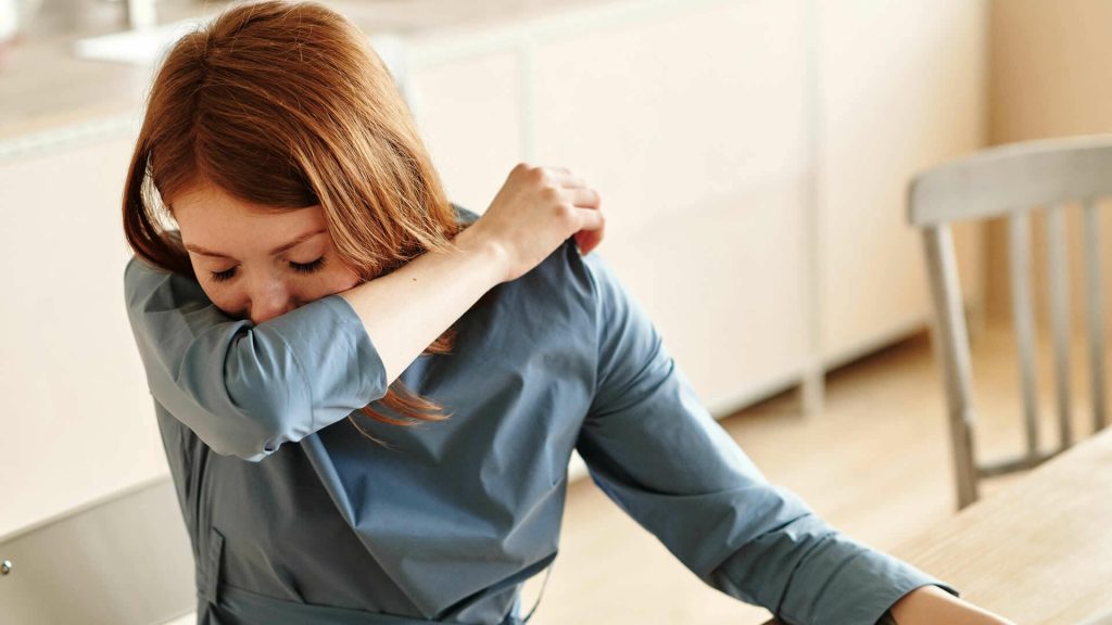 A woman with red hair in a blue shirt sneezes into her elbow while sitting at a table in the medical center.