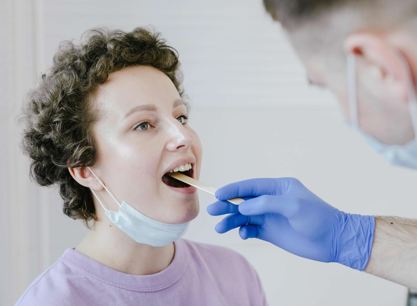 A medical professional in a blue surgical mask and gloves examines a patient wearing a lavender shirt. 