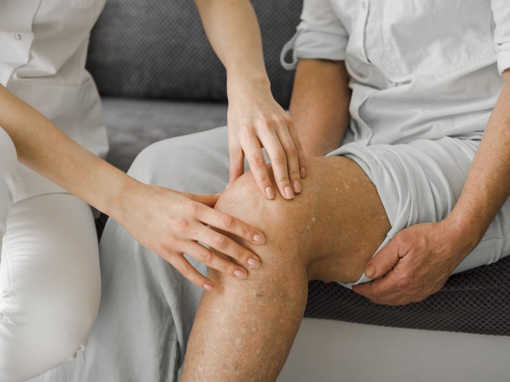 A healthcare professional examines a seated person's knee, with both hands gently touching the patient's knee area.