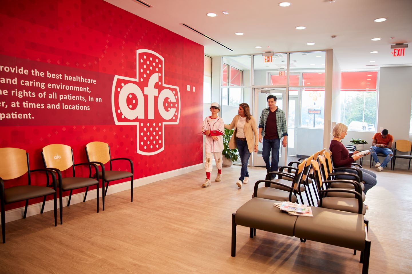 Waiting Area in a Healthcare Facility: The image depicts an interior space, likely a waiting area in a healthcare facility. Rows of chairs are arranged along the left wall, and there’s a red accent wall with partially visible white text. People are either seated or walking, and large windows allow natural light into the room.Waiting Area in a Healthcare Facility: The image depicts an interior space, likely a waiting area in a healthcare facility. Rows of chairs are arranged along the left wall, and there’s a red accent wall with partially visible white text. People are either seated or walking, and large windows allow natural light into the room.