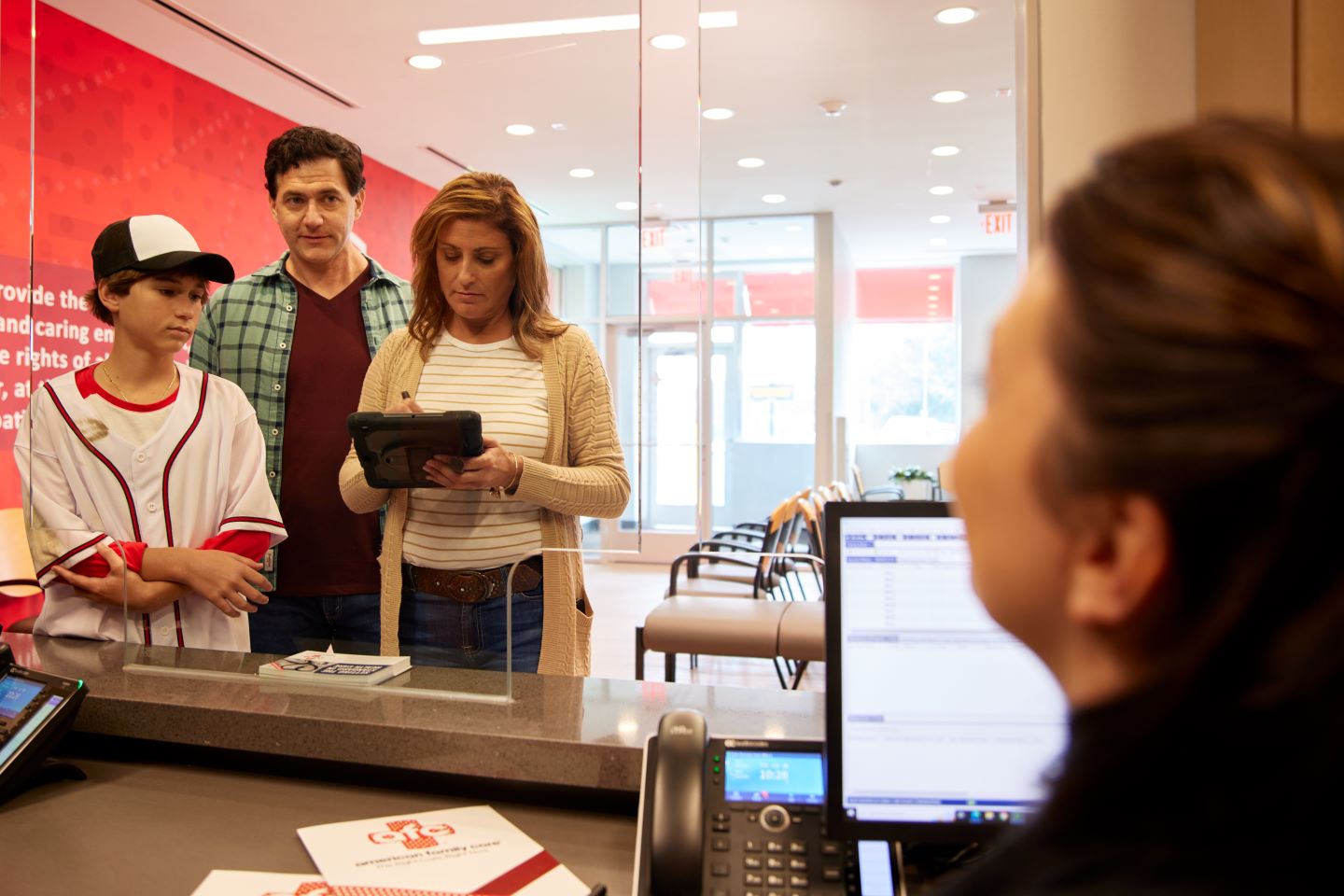 A woman signs a form on a tablet at a reception desk, while a man and a child, who is wearing a baseball uniform, stand beside her. Another person, the receptionist, is seated behind the desk. The setting is a medical office or clinic.